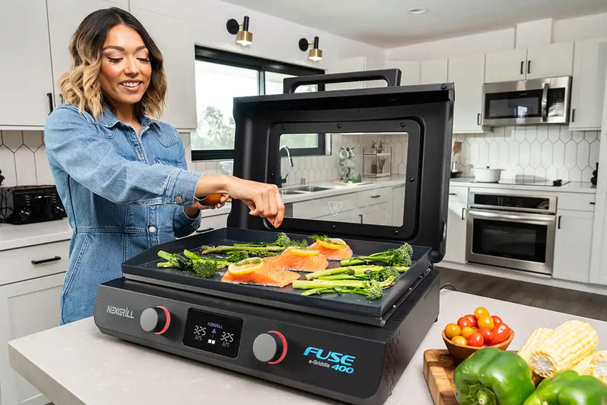 A woman cooks salmon and vegetables on an electric indoor grill in a modern kitchen. Fresh ingredients and bright vegetables are on the counter nearby.