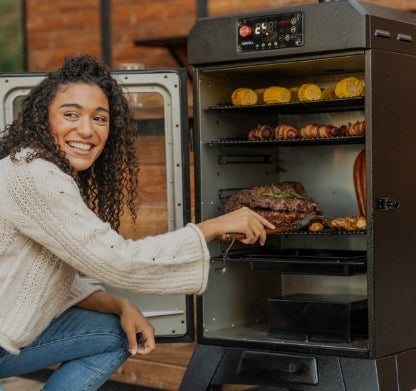 Smiling woman kneeling by an open electric smoker filled with grilled meat, corn on the cob, and vegetables. She is pointing at the food inside while wearing a cream sweater and jeans.