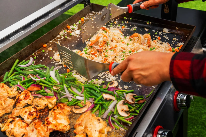 Hands cooking fried rice, vegetables, and grilled chicken on a large outdoor flat-top grill. Green beans, mushrooms, and onions are visible among the ingredients.